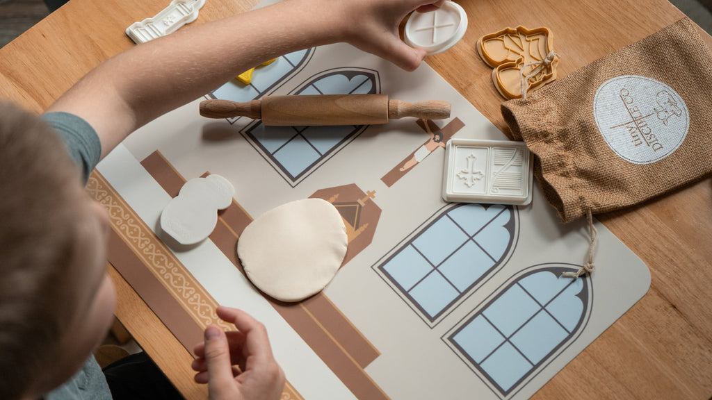A child's hands are shown picking up a plastic play dough mold in the shape of a host. there is also a rolled out white piece of play dough laying on top of a silicone mat that depicts the image of an altar table. 