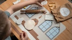 A child's hands are shown picking up a plastic play dough mold in the shape of a host. there is also a rolled out white piece of play dough laying on top of a silicone mat that depicts the image of an altar table. 
