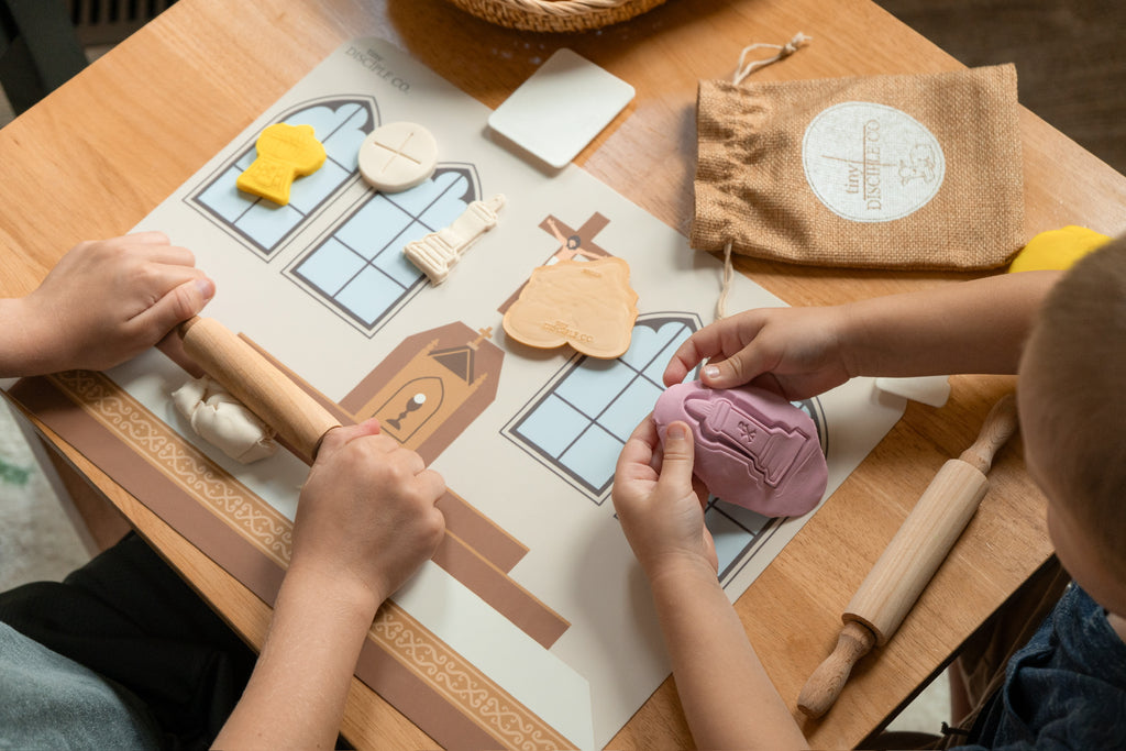 A top-down view of children's hands playing with a  silicone mat the plastic play dough showing specifically the bells, candle, thurible and missile molds and play dough.