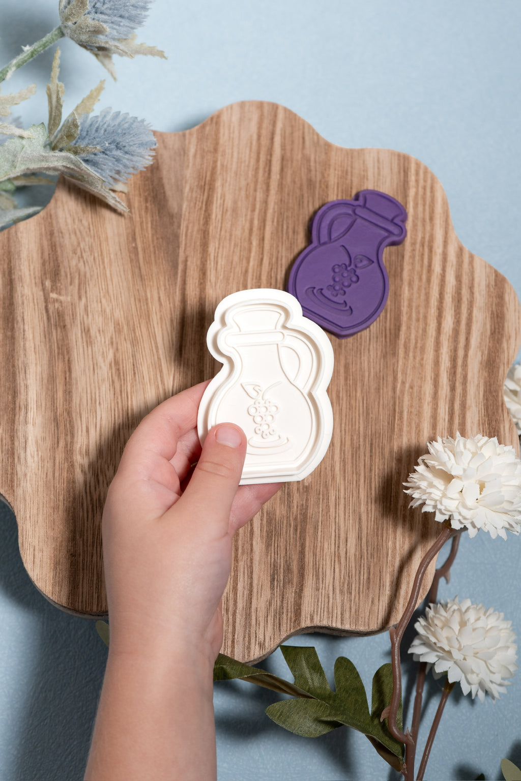 A hand holds a white silicone mold shaped like a wine decanter, with a purple matching play dough cut out resting on a wooden board against a light blue background with decorative flowers.
