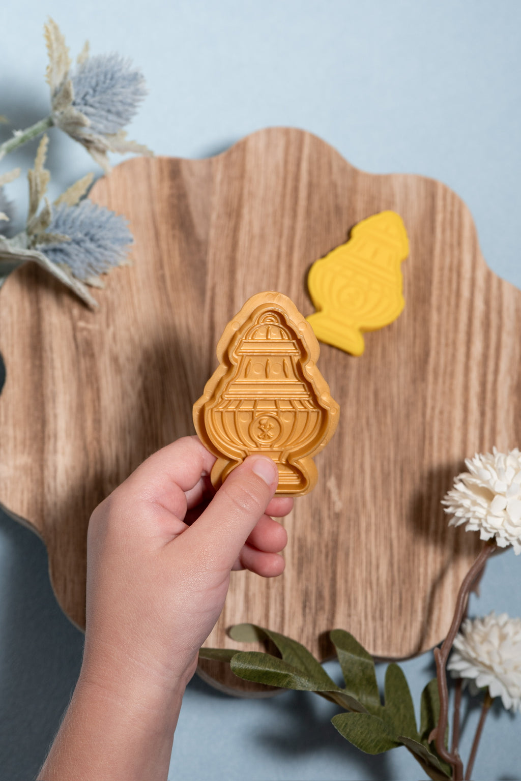 A hand holds a gold thurible play dough mold and a matching golden-thurible cut out is visible in the background on a scalloped wooden board, set against a light blue backdrop with decorative dried flowers.