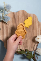 A hand holds a gold thurible play dough mold and a matching golden-thurible cut out is visible in the background on a scalloped wooden board, set against a light blue backdrop with decorative dried flowers.