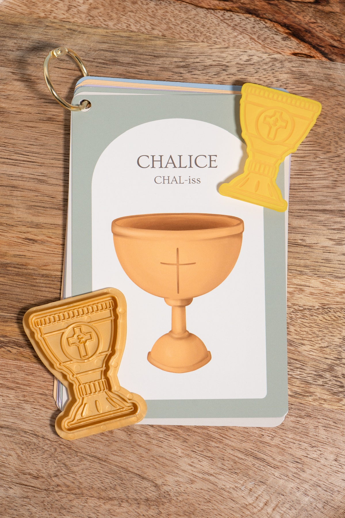 A small printed card titled "CHALICE" with an illustration of a chalice, placed on a wooden surface alongside two small yellow chalice-shaped pieces, one made of play dough and one that is a plastic cut out mold.