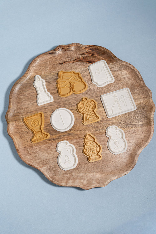 A set of plastic play dough molds that press out images of items used during a catholic mass. these items include a candle, bells, purificatory, chalice, host, thurible, missal, water decanter, wine decanter and ciborium. they are displayed on a wooden circle plate. 