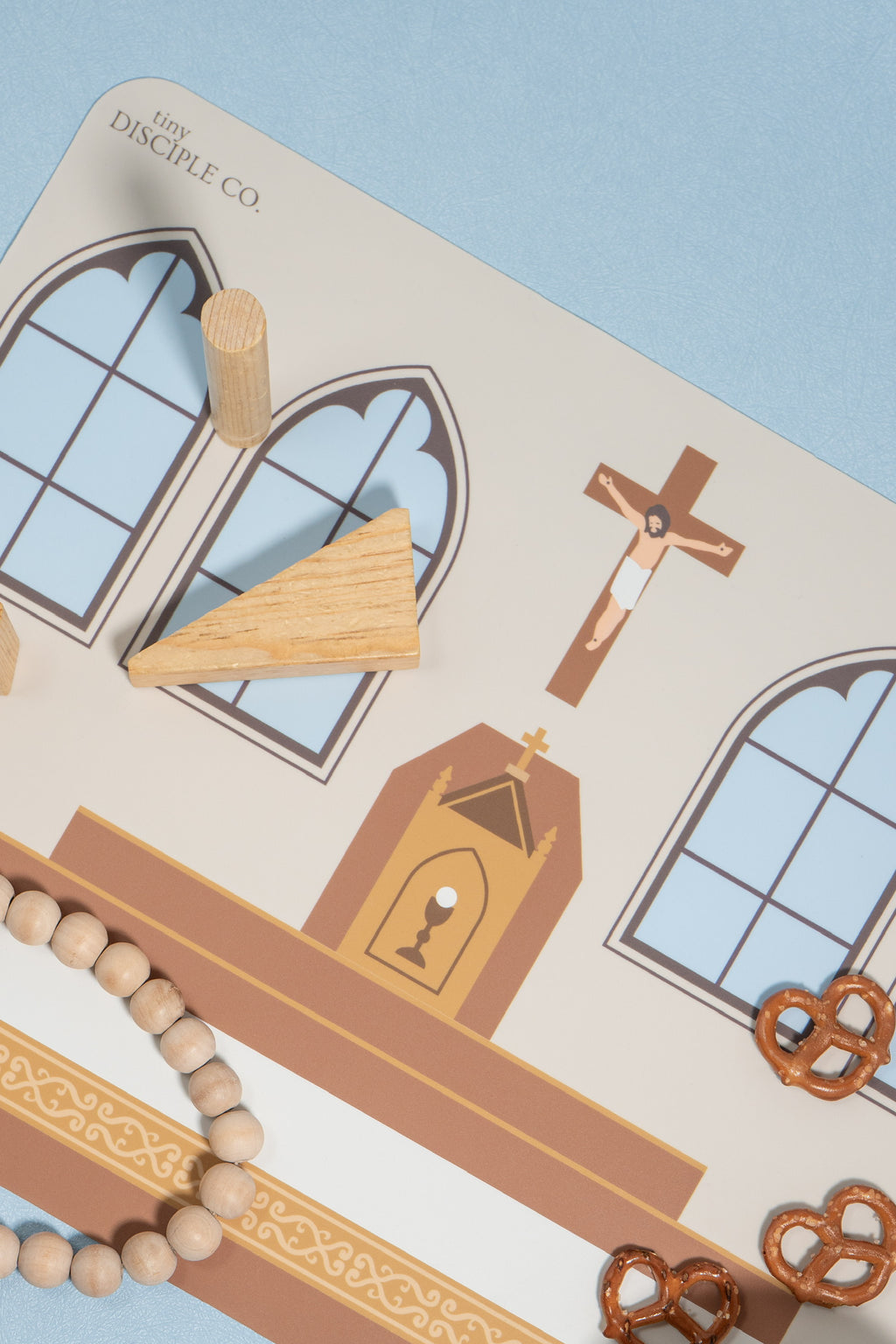 A close-up of a silicone mat with an image of a Catholic Church altar table. This table has wooden pieces, including a triangular piece, a wooden cross, and a beaded bracelet, arranged on a light blue background.