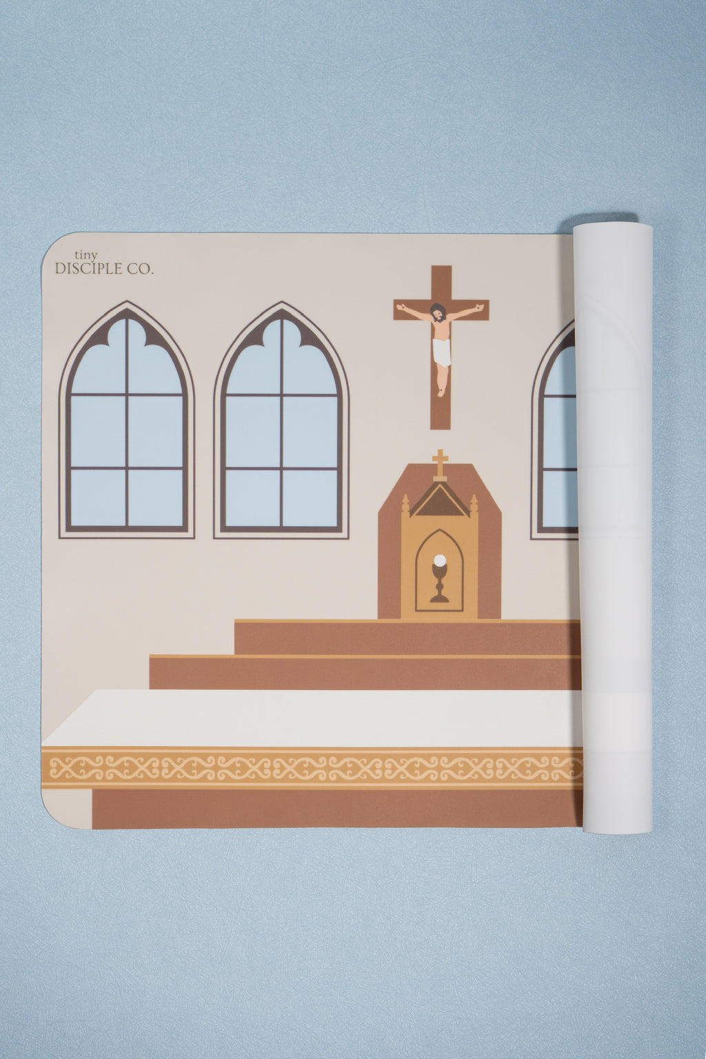 A rolled-up printed mat showing a catholic church altar interior with arched windows, an altar, and a crucifix, photographed on a light blue background. The printed design includes the text 'The Disciple Co.