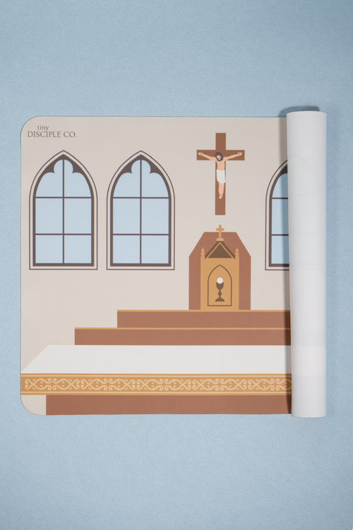 A rolled-up printed mat showing a catholic church altar interior with arched windows, an altar, and a crucifix, photographed on a light blue background. The printed design includes the text 'The Disciple Co.